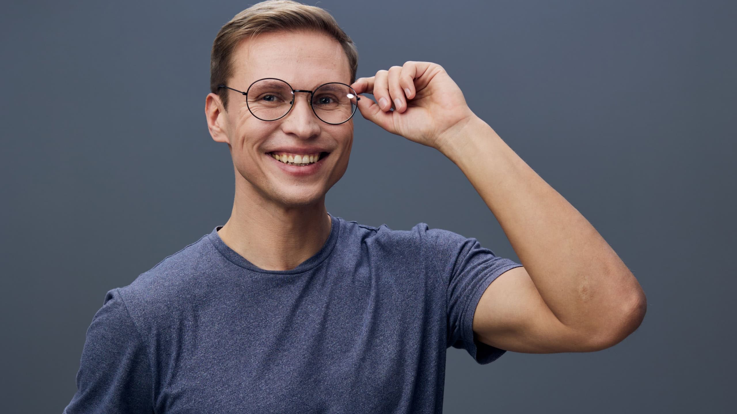 Confident young man wearing glasses and casual grey t-shirt smiles at camera on plain grey background studio portrait. People lifestyle concept
