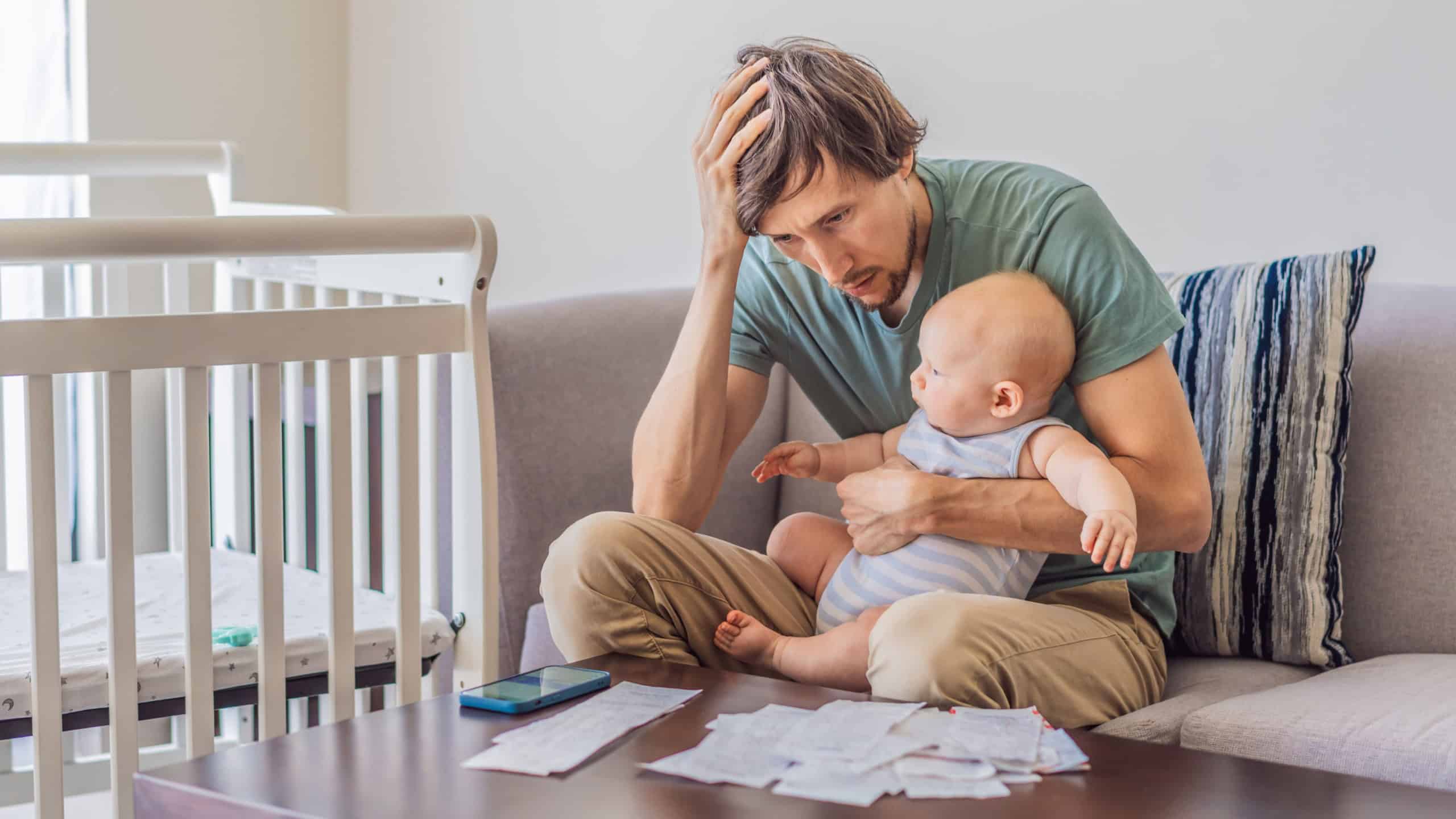 Father sitting on sofa holding baby while calculating monthly expenses with receipts at home. Stressed dad managing budget and childcare. Financial stress, parenthood and real life concept