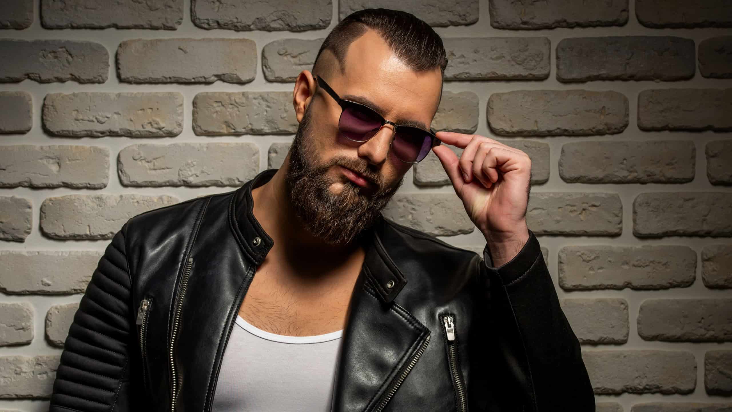 Stylish bearded man in sunglasses and leather jacket posing against a brick wall.
