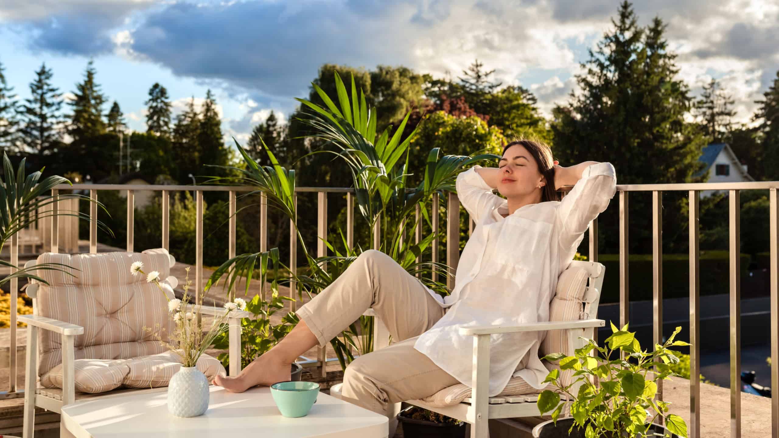 Beautiful young girl relaxing and enjoying sun while having tea, sitting at balcony at sunlight at summer. Slow living, tranquil moment, mental health. Backyard terrace vacation.