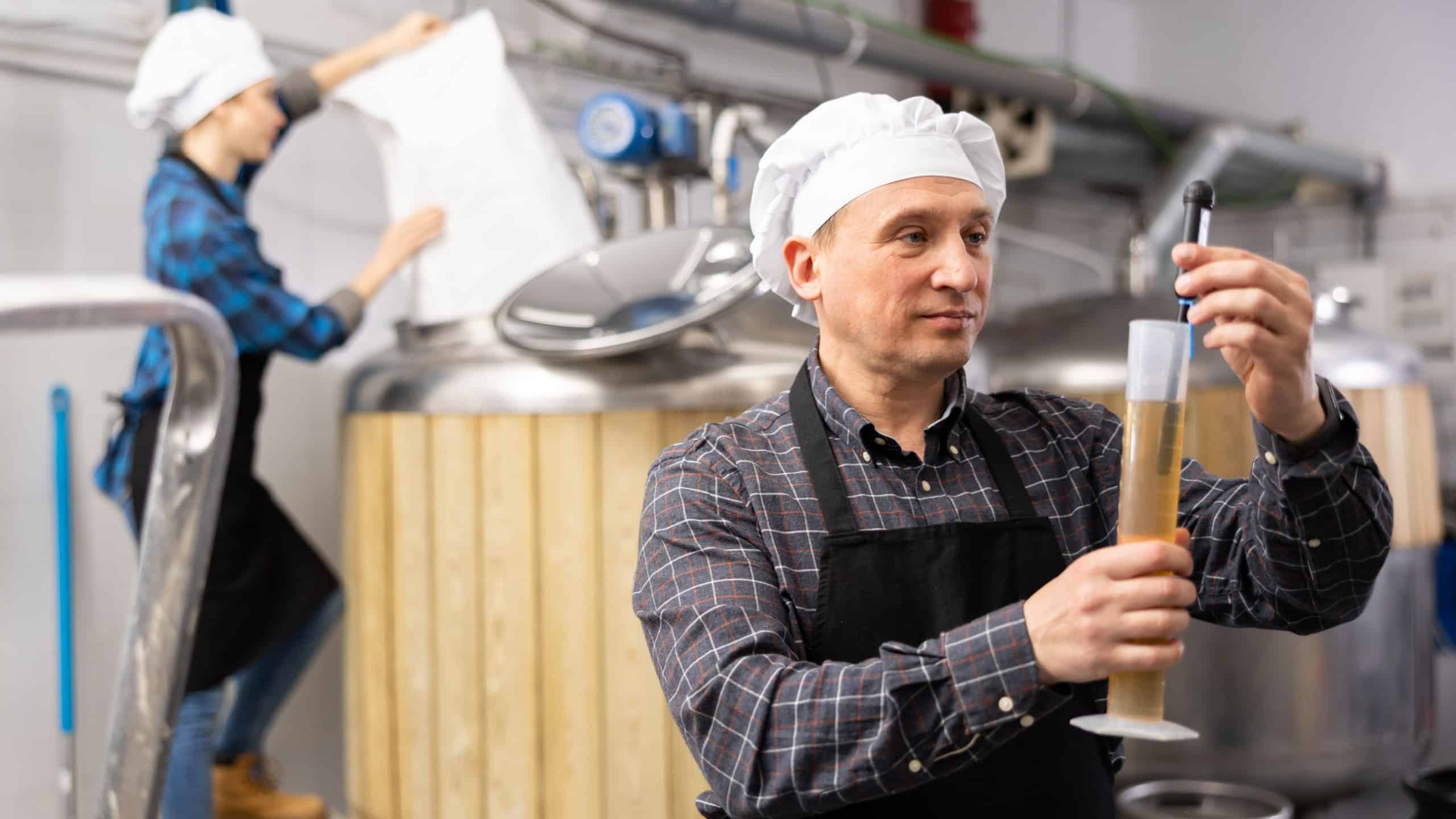 Professional brewmaster controlling craft beer production process in brewery, measuring beverage density with digital hydrometer