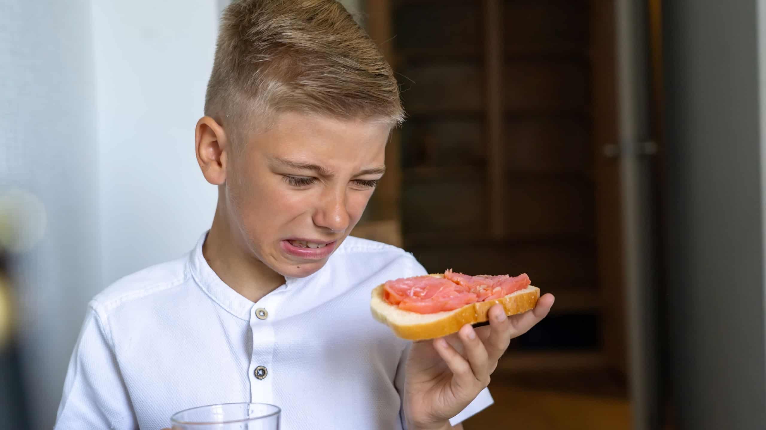 Upset disgruntled teenager grimaces and holds a fish sandwich at breakfast in the kitchen