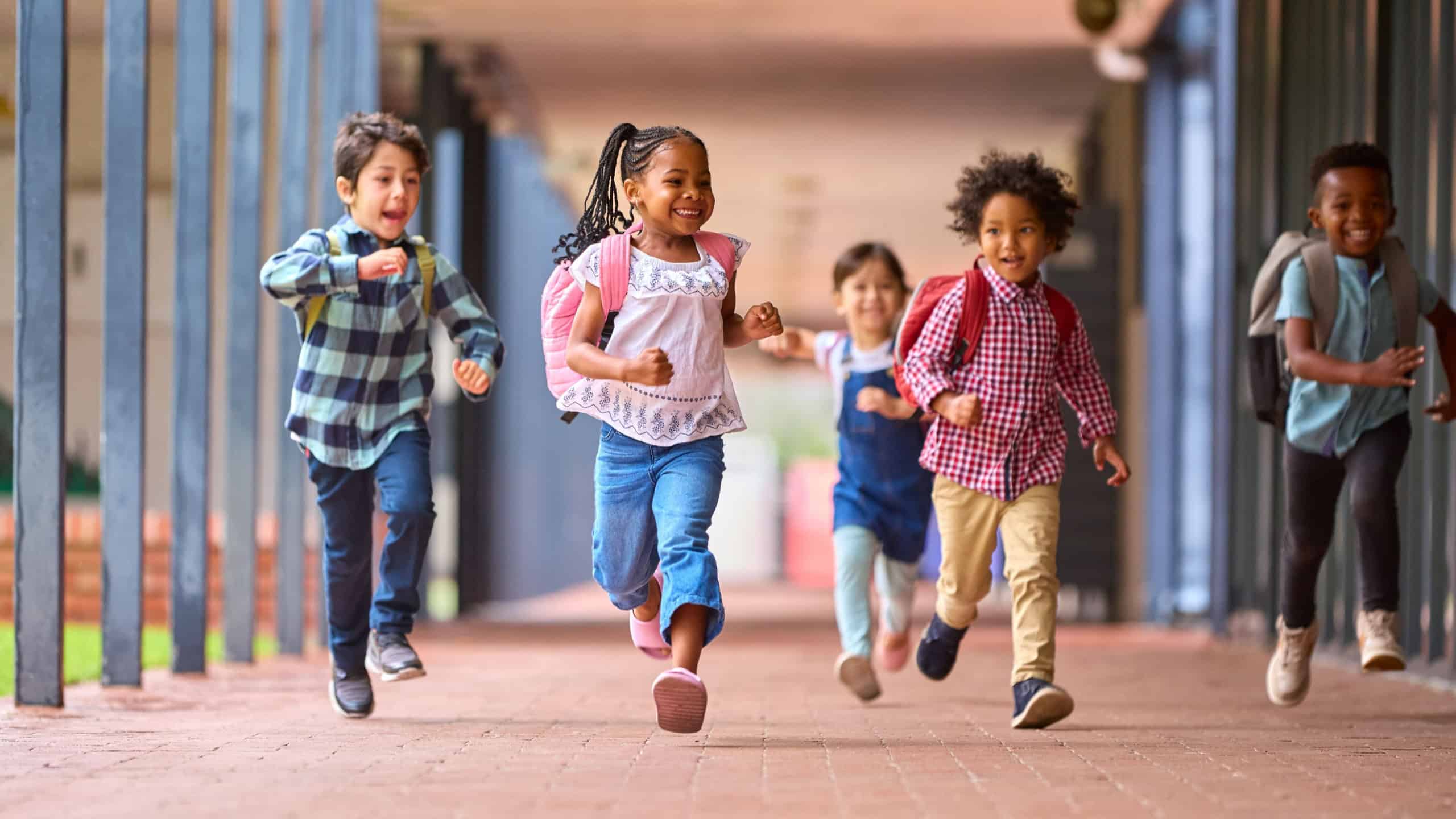 Group Of Multi-Cultural Elementary School Pupils Running Along Walkway Outdoors At School
