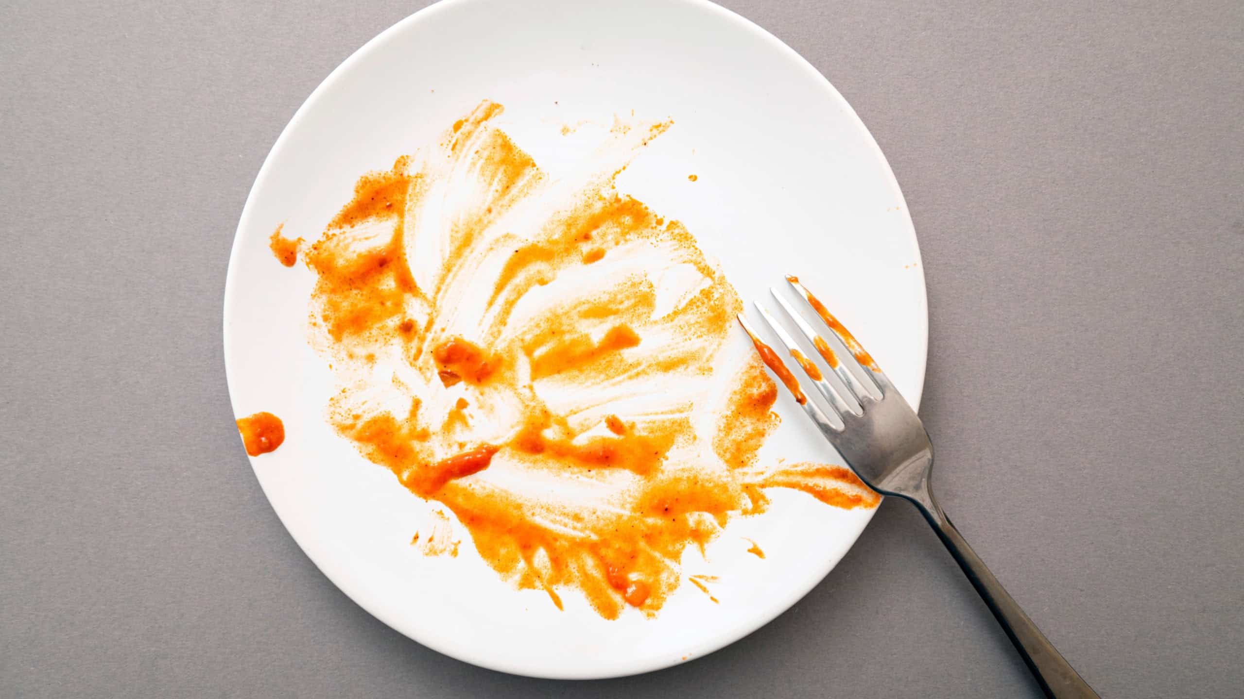 Dirty white ceramic plate with tomato sauce stains, food leftovers and fork, top view