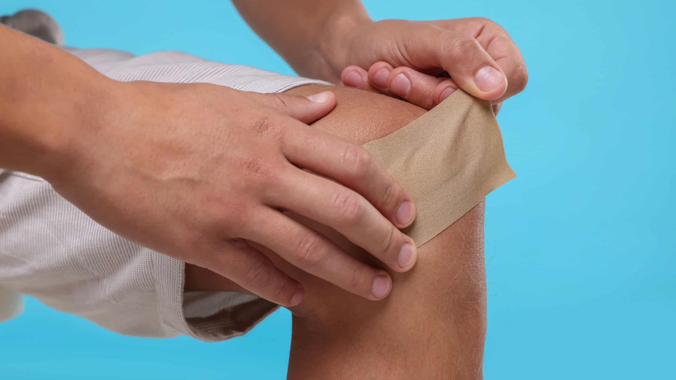 Man putting sticking plaster onto knee on light blue background, closeup