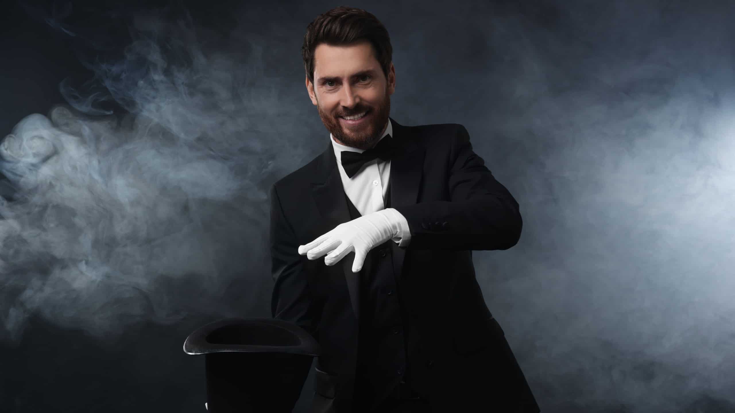 Happy magician showing magic trick with top hat in smoke on dark background