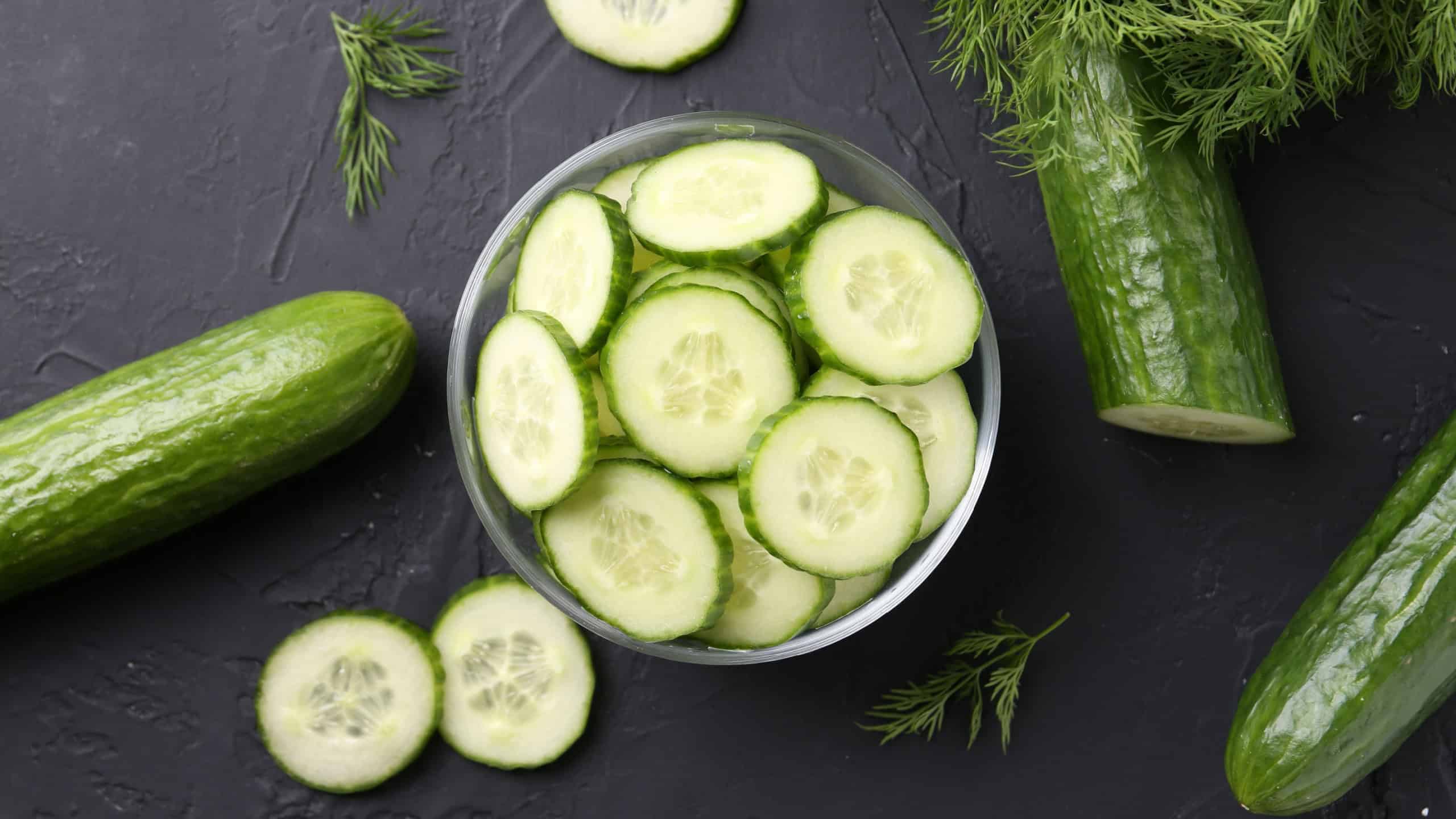 Cut cucumber in glass bowl, fresh vegetables and dill on dark gray textured table, flat lay