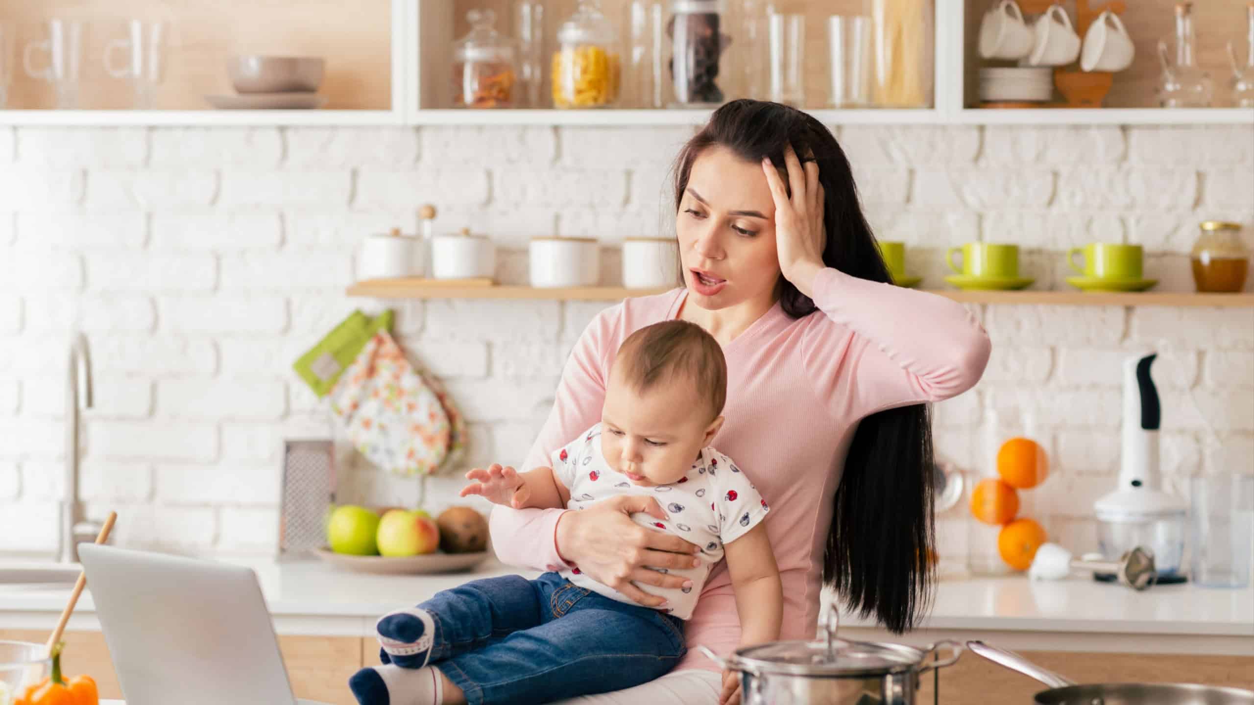 A mother tries to manage her remote work on a laptop while holding her baby in a modern kitchen, highlighting the challenges of multitasking with household duties and childcare responsibilities.