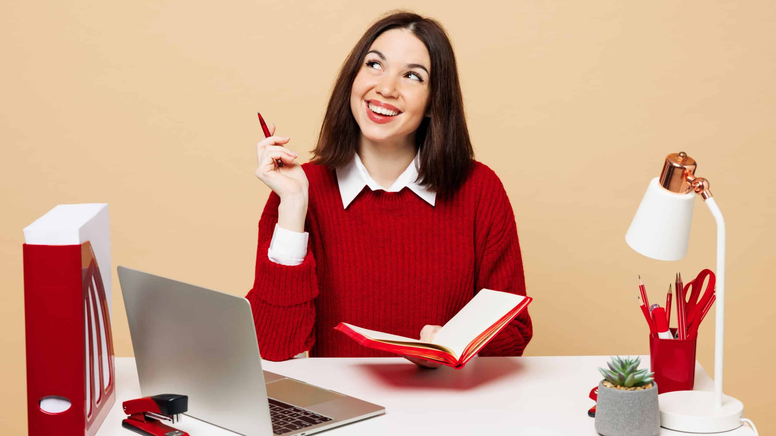 Young minded employee business woman wear red sweater shirt sit work at office desk with pc laptop hold notebook pen look aside isolated on plain pastel beige background. Achievement career concept