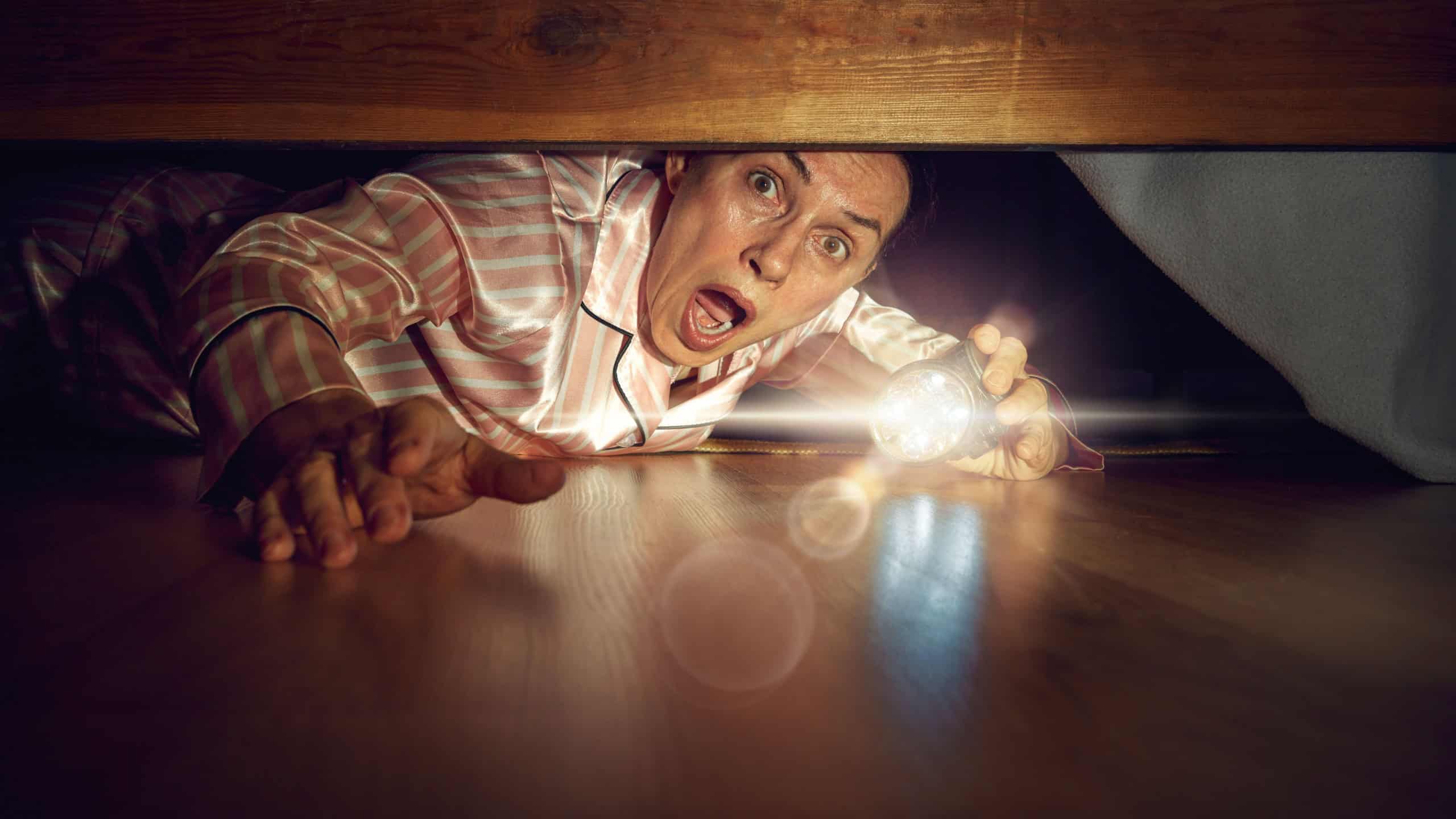 Senior woman with emotional face, lying on floor and looking under bed with flashlight, showing emotions of fear. Spooky season., Concept of secrets, human emotions