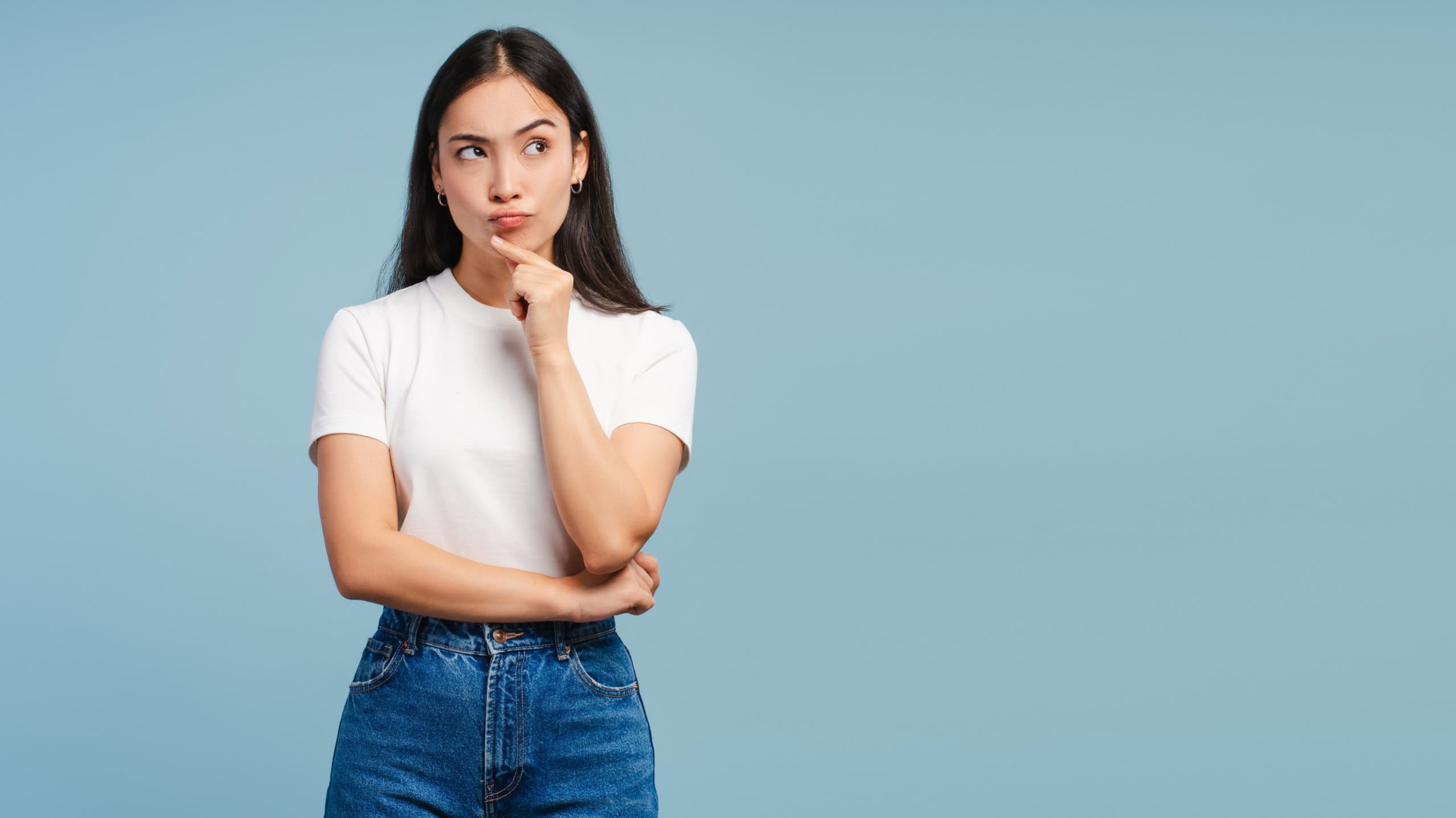 Pensive young Asian woman thinking with finger on chin posing on blue background copy space. Advertisement concept