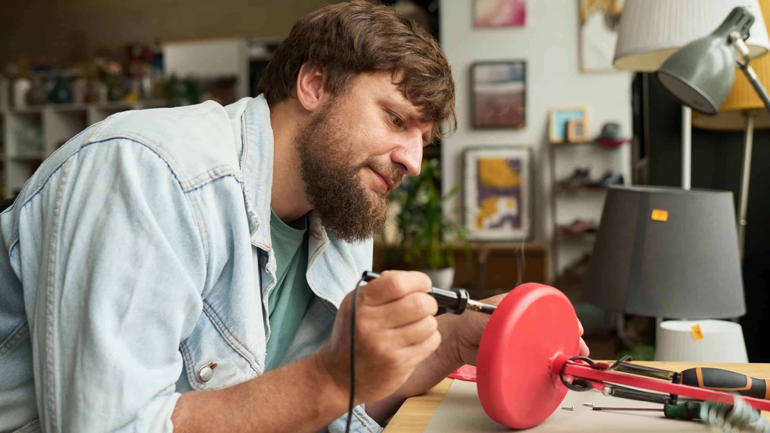 Bearded man bending over table and soldering bottom part of red secondhand electric lamp while repairing it in thrift shop