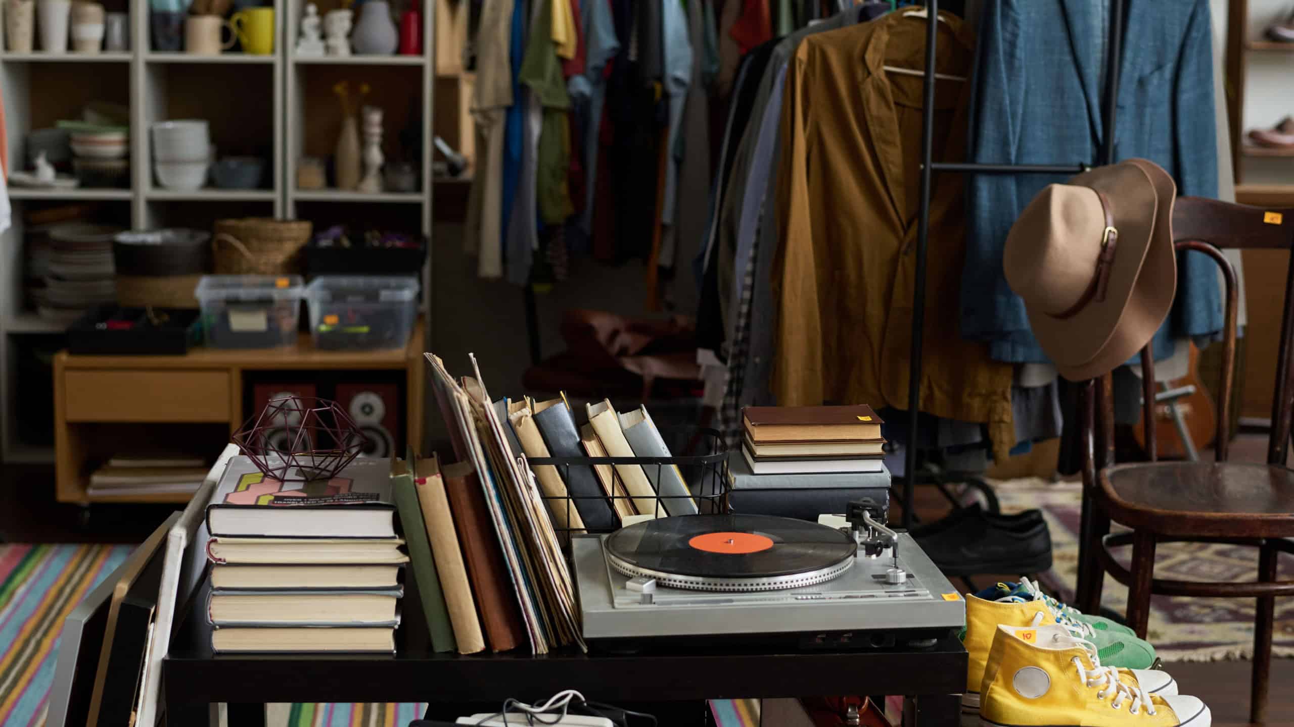 Several stacks of books, stereo record player on table standing by suitcases with sports shoes on top against racks with attire
