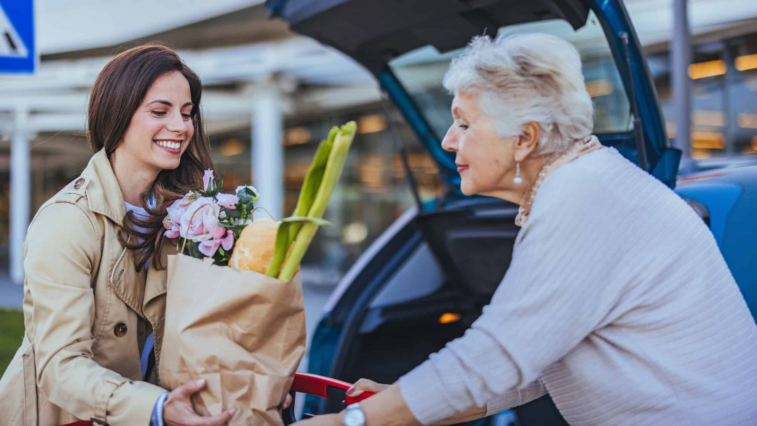 A young woman assists an elderly lady in unloading groceries from a car. The scene conveys a sense of community, assistance, and friendship, highlighting the joy of helping others.