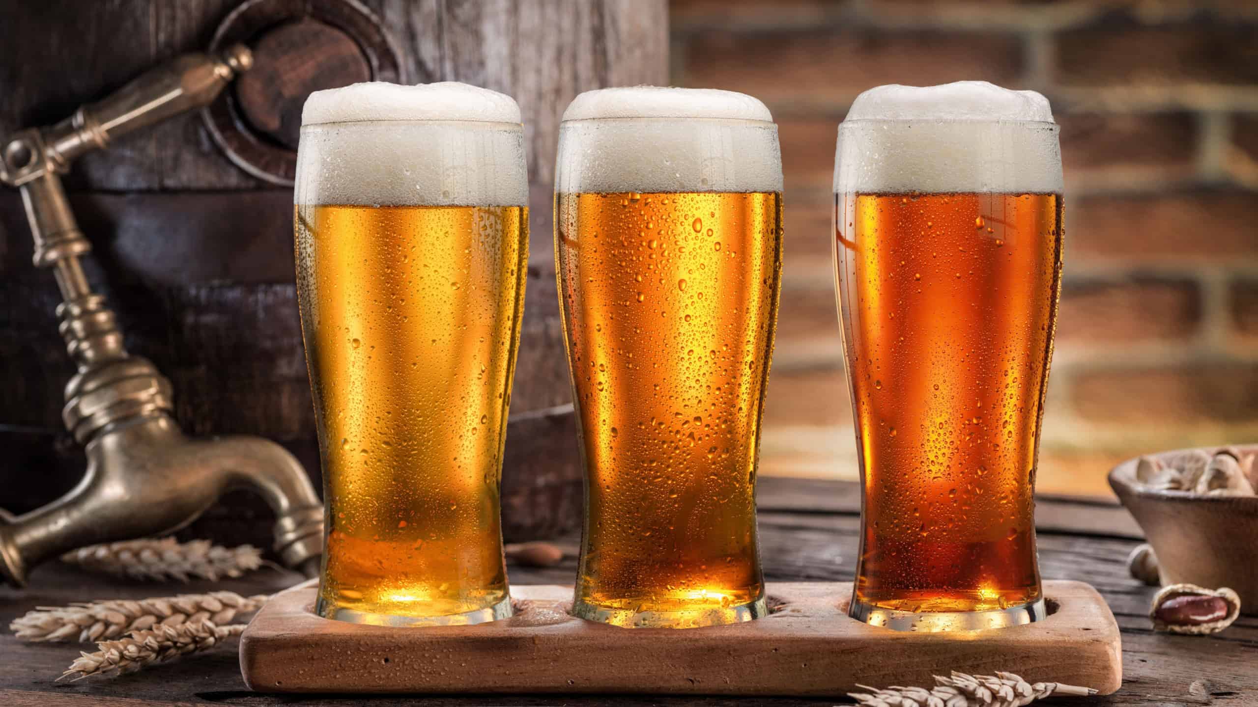 Three glasses of chilled beer near beer cask on old wooden table and brick wall at the background. 