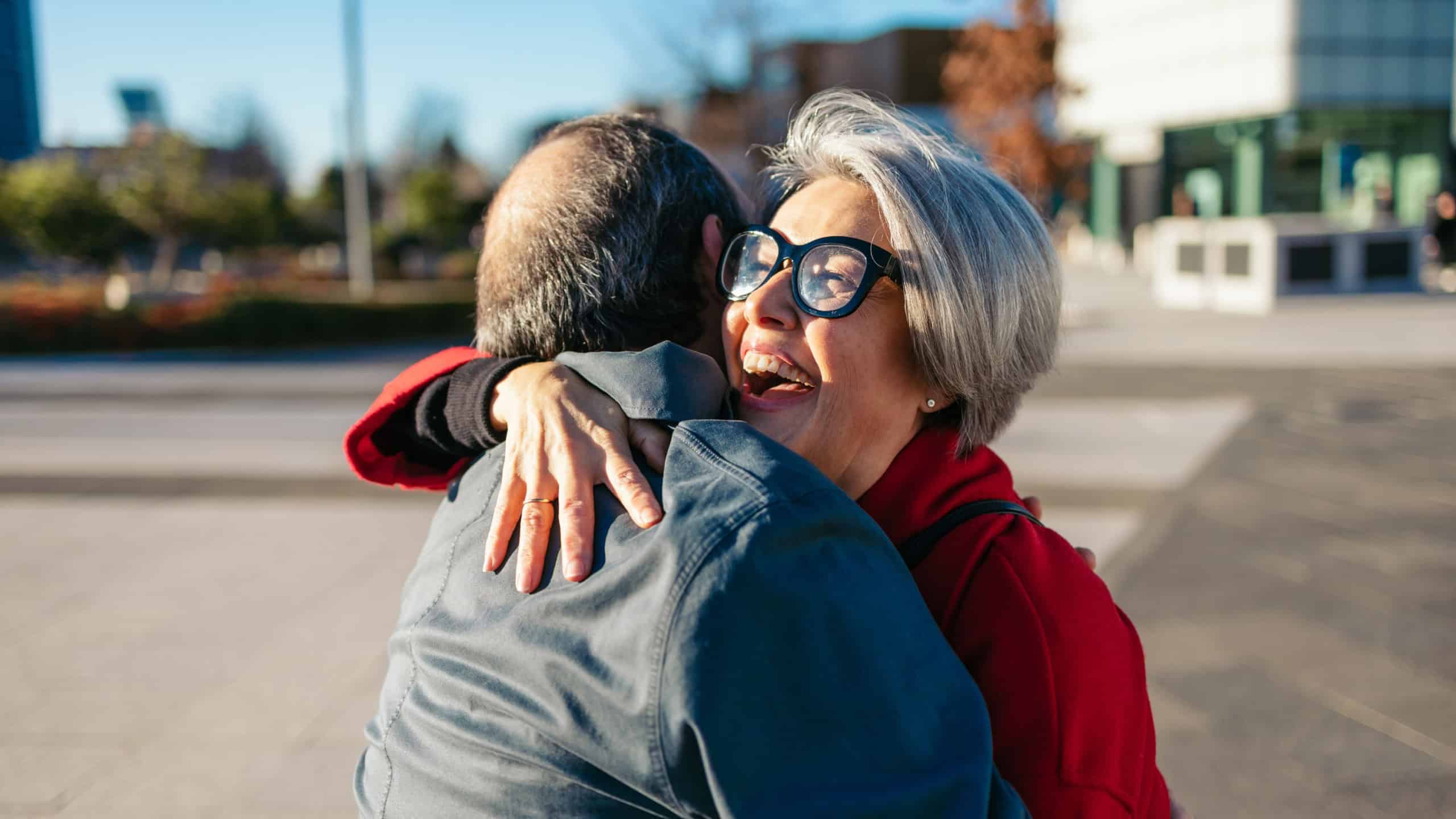 Mature woman wearing bright red coat hugging smiling friend on city street corner