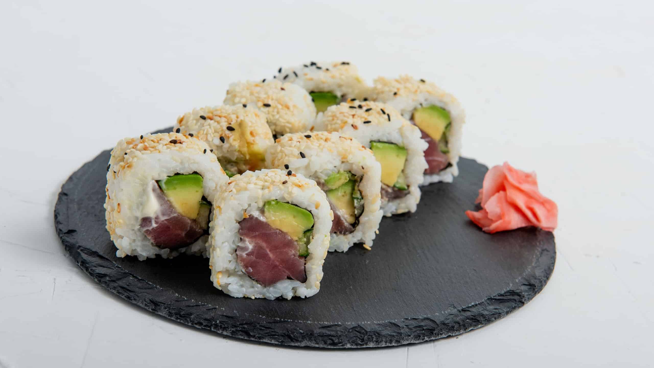 Close-up of sushi California rolls with tuna, avocado, and sesame seeds, served on a black slate plate against a white isolated background