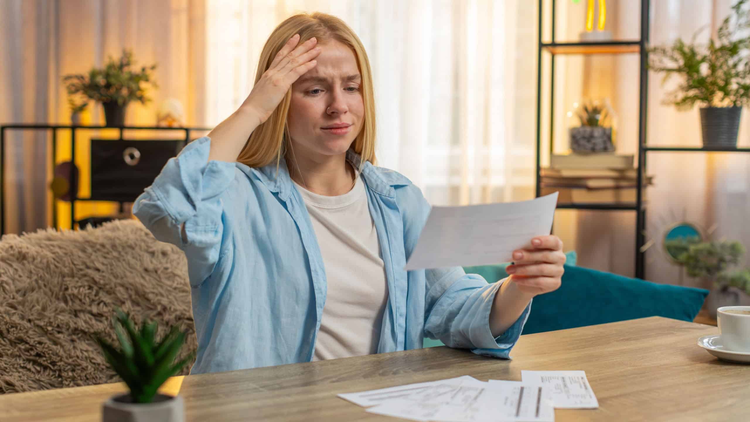 Young woman checks statement at home, eyes widening with alarm over unexpected expense. Girl sits at table sets paper aside, revealing tense realization of higher than normal cost of utility services