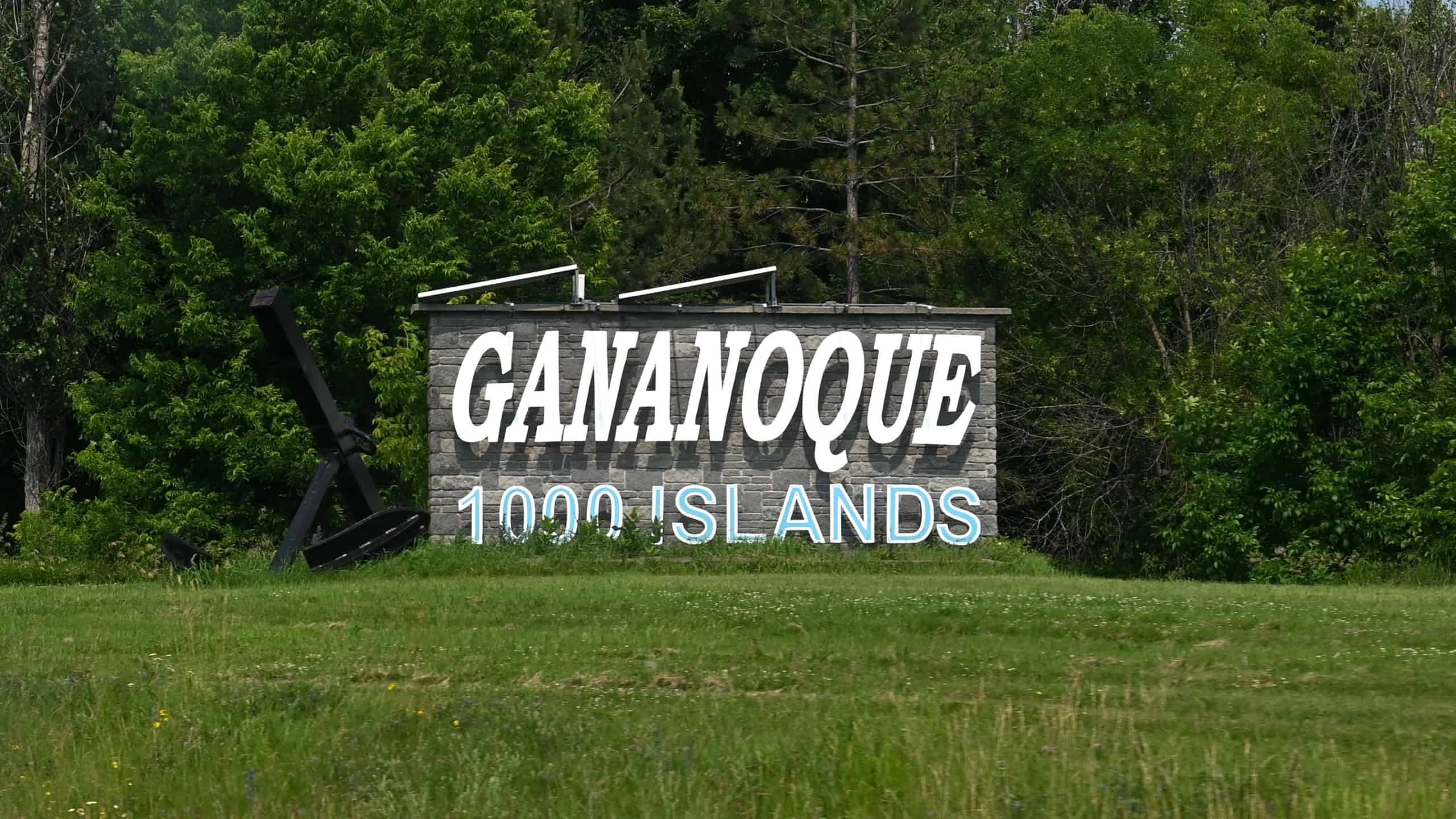 Gananoque, ON, Canada - September 16, 2025: A welcome sign for the town of Gananoque stands on a grassy hill next to a road on a sunny day.