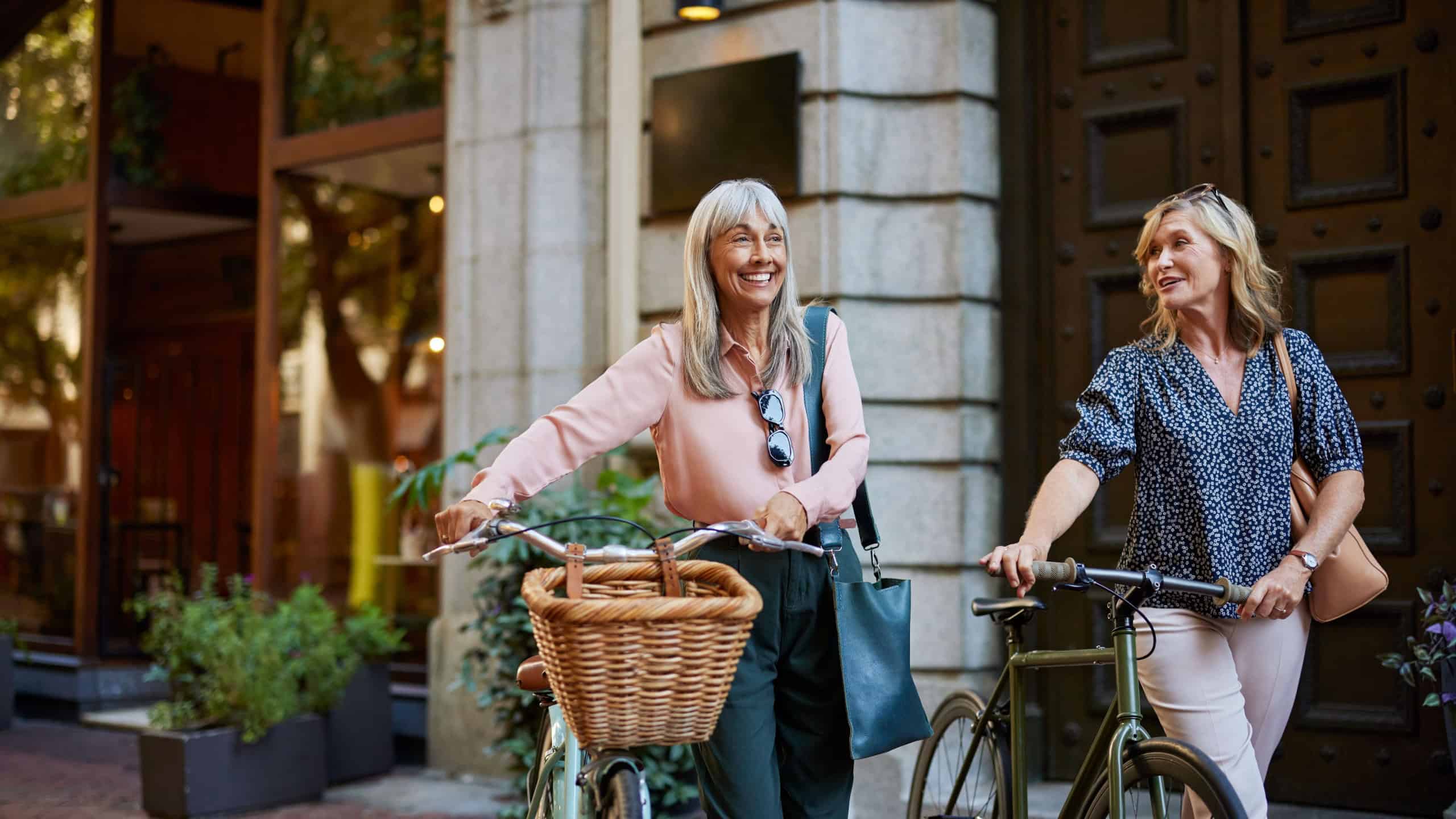 Two older happy women smiling and pushing their bicycles outdoors. Mature friends enjoy a casual walk with their bikes on a city street. Happy retired healthy women walking with bicycles in street.