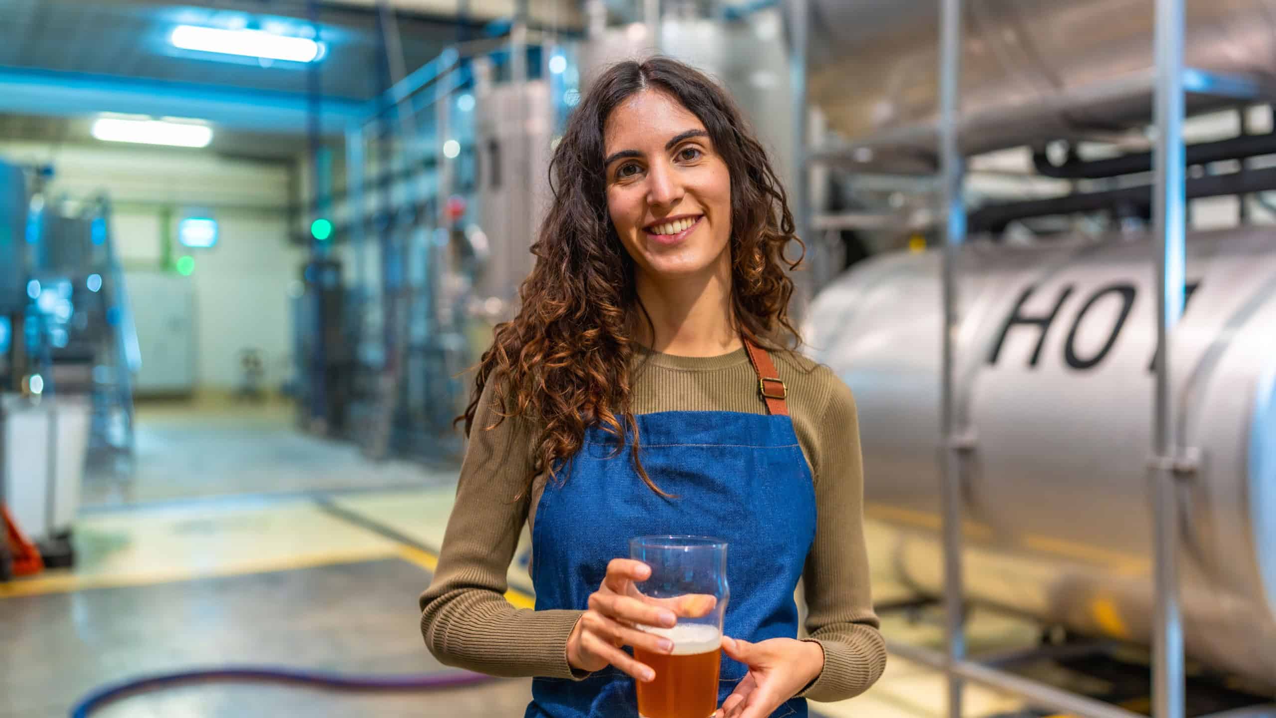 Woman brewer standing in modern brewery, smiling and holding a glass of craft beer for quality tasting, representing small batch production, craftsmanship and confident ownership