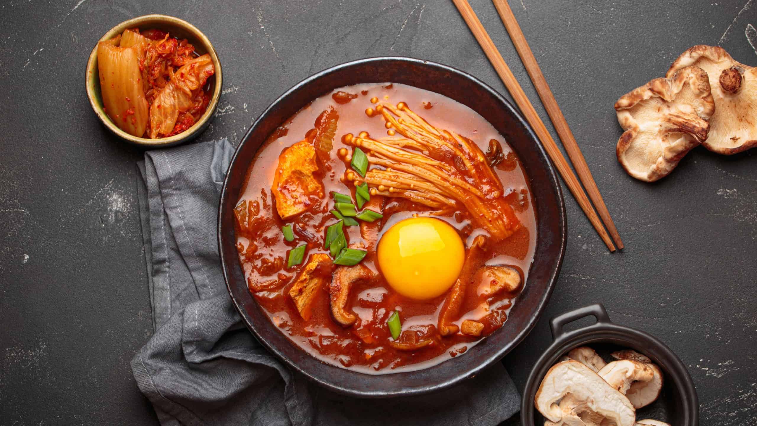 Traditional Korean kimchi jjigae hot soup or stew with egg yolk in black rustic bowl on dark background top view