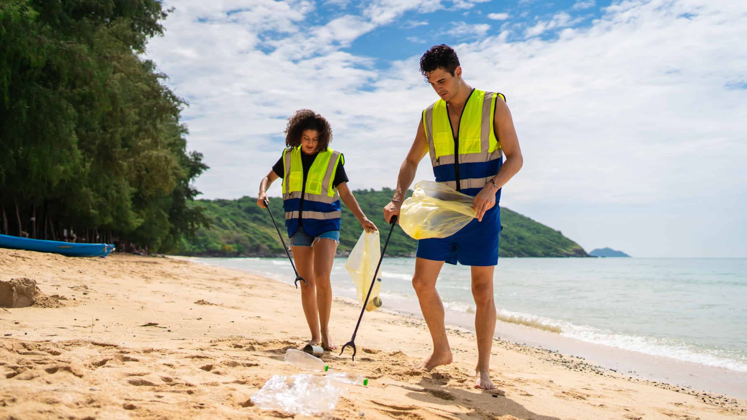 Two volunteers collecting plastic bottles and trash on a sandy beach. Caucasian man and Latino woman wearing vest, work together to clean the beach and protect environment, with ocean in background.