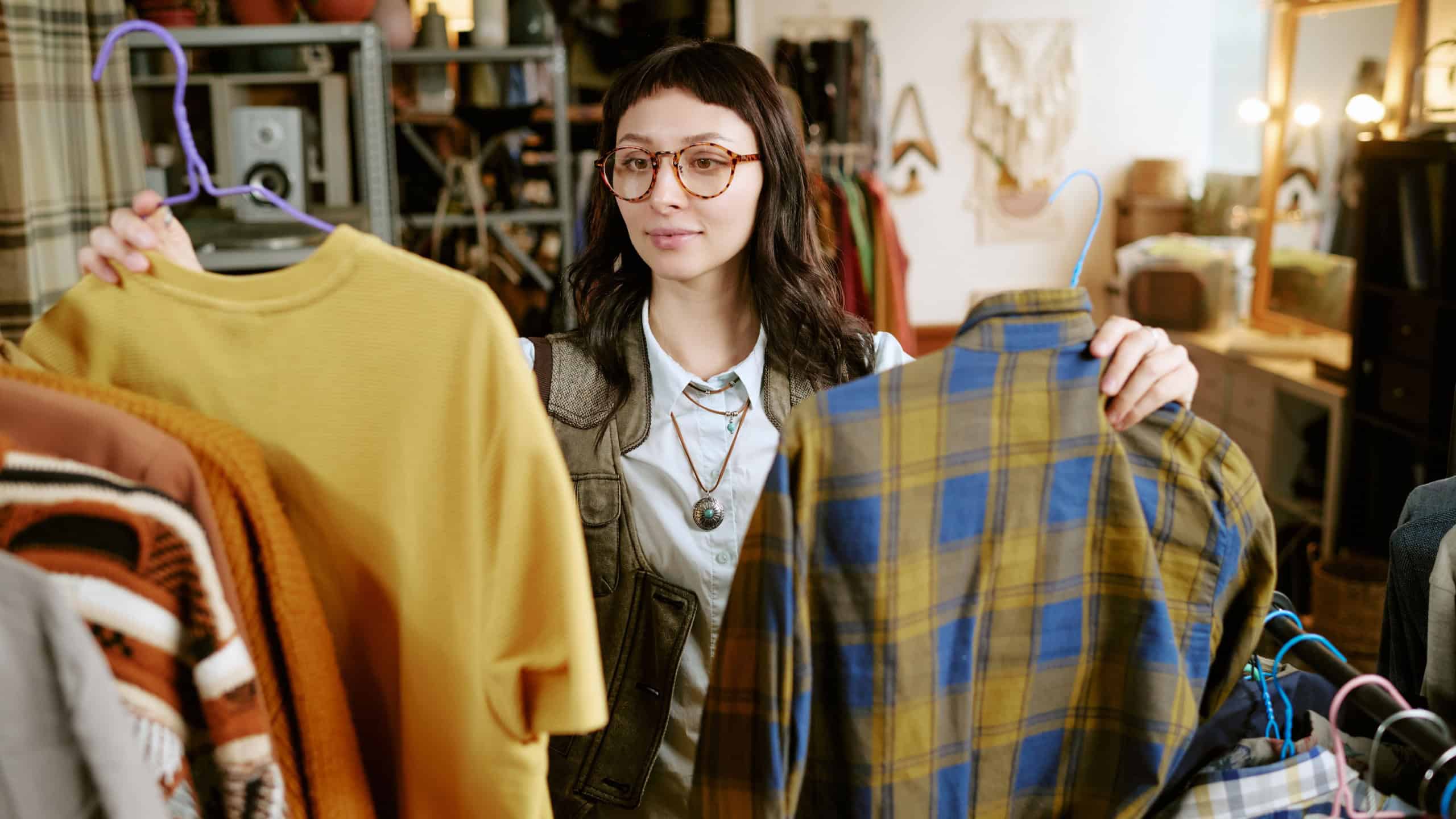 Young adult biracial woman browsing second hand clothing store, holding two shirts on hangers and comparing options, surrounded by racks of used garments and accessories