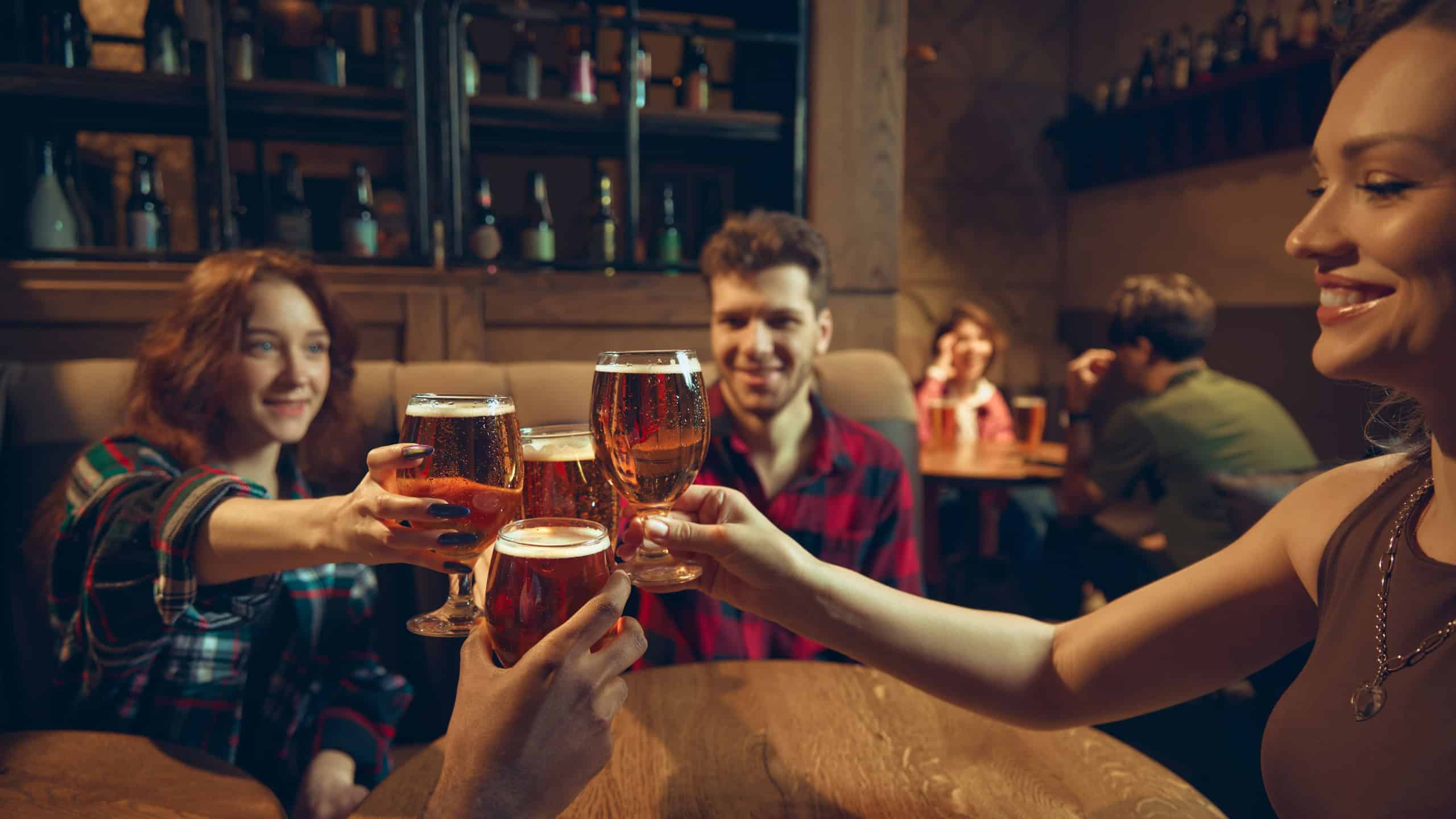 Group raising beer glasses around table in warm pub interior. Concept of nightlife promotion, bar culture visuals, brewery campaigns, social get-togethers, and relaxed weekend celebrations.