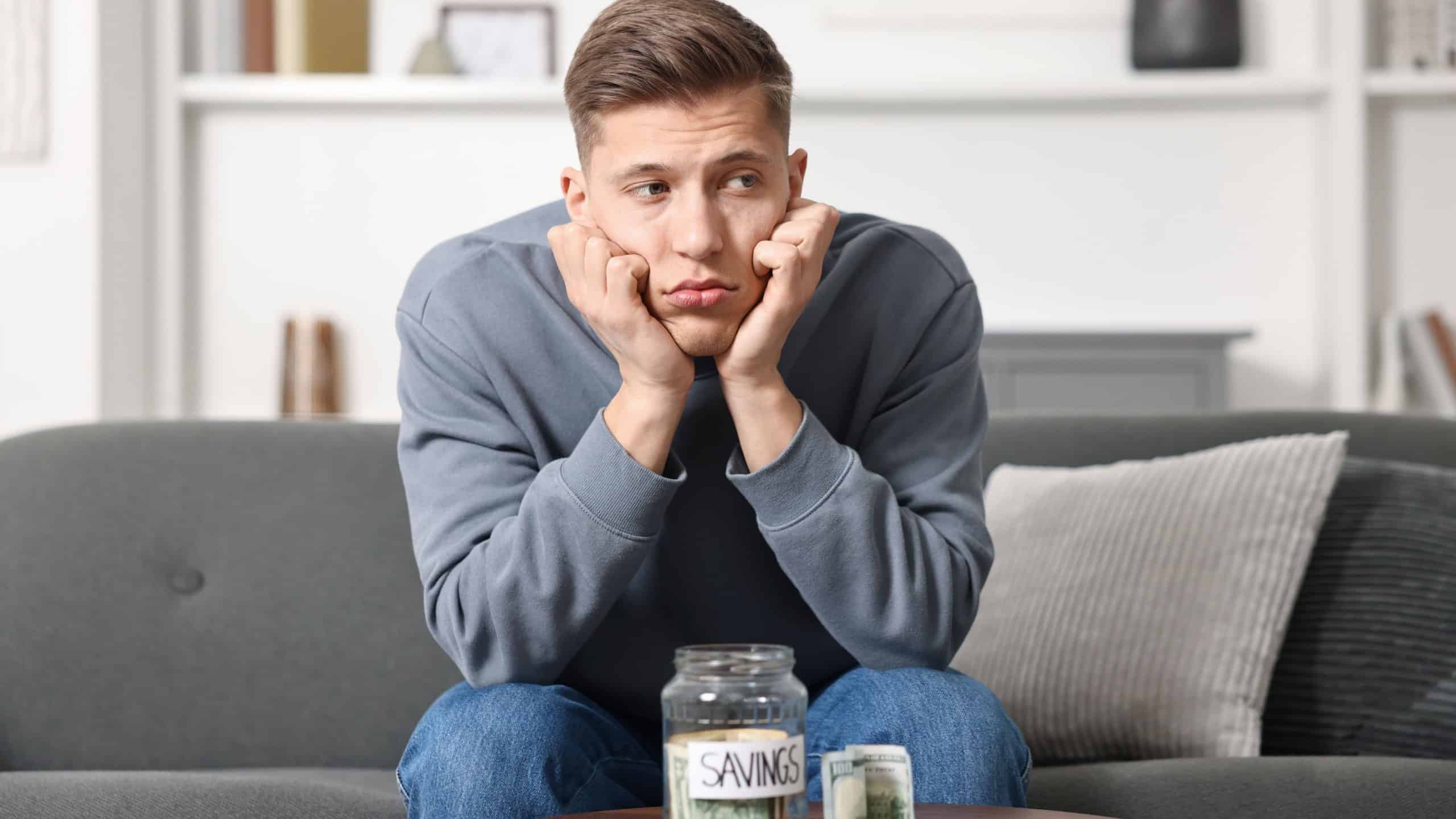 Sad man with savings jar and money at wooden table indoors