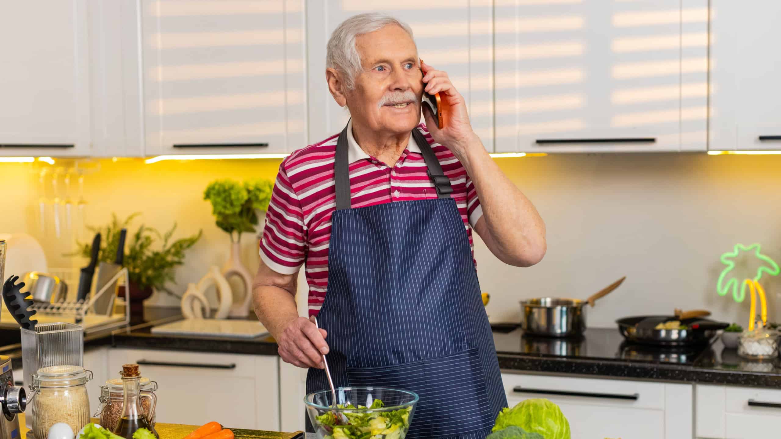 Senior old man smiles while mixing fresh salad in home kitchen talking on smartphone about diet menu daily. Elderly grandfather enjoys healthy eating lifestyle during call planning weight loss meal.