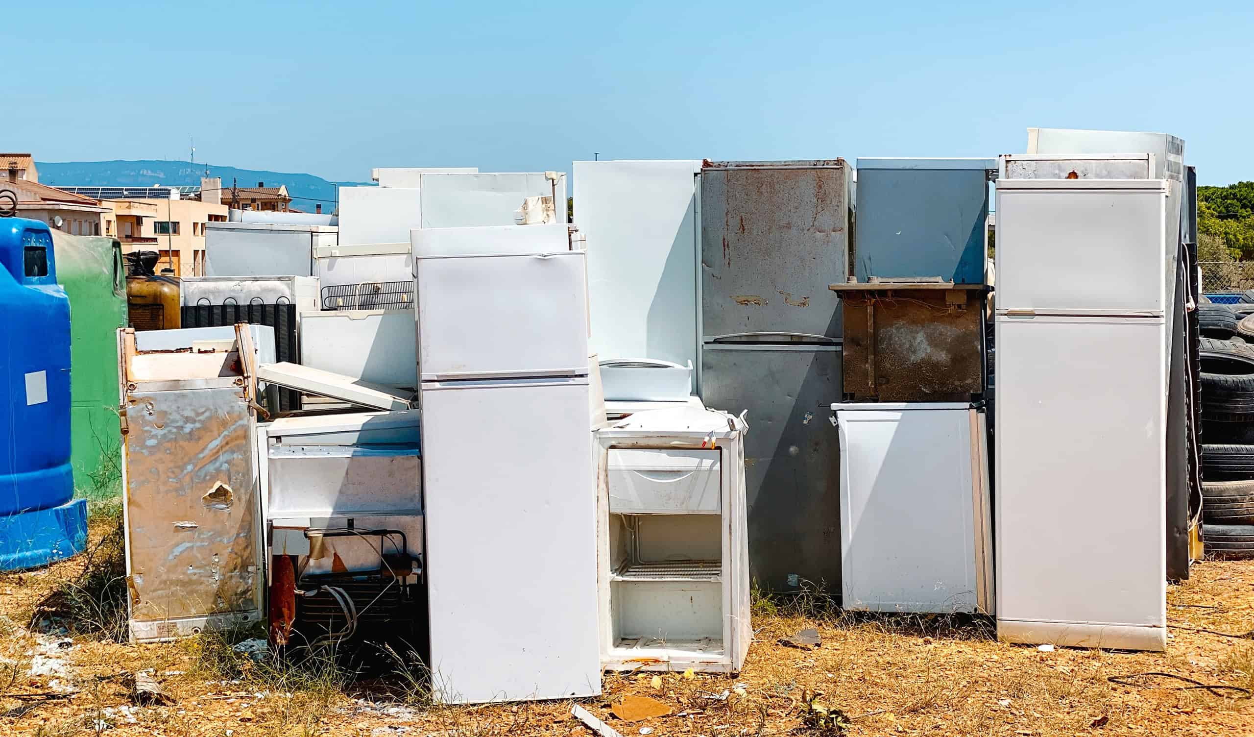 an outdoor recycling yard section with a pile of discarded refrigerators and freezers stacked outdoors under the sun showing metal rust and old appliances waiting for disposal