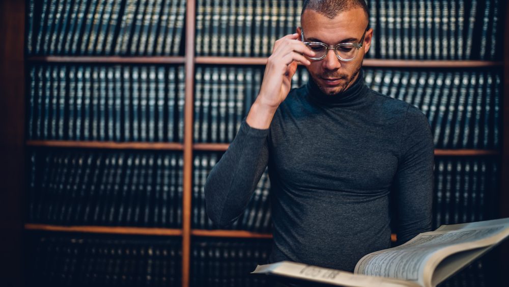 Serious male scientist in eyeglasses reading literature spending time on autodidact in public library, clever journalist making researchers for working task standing near bookshelves collections