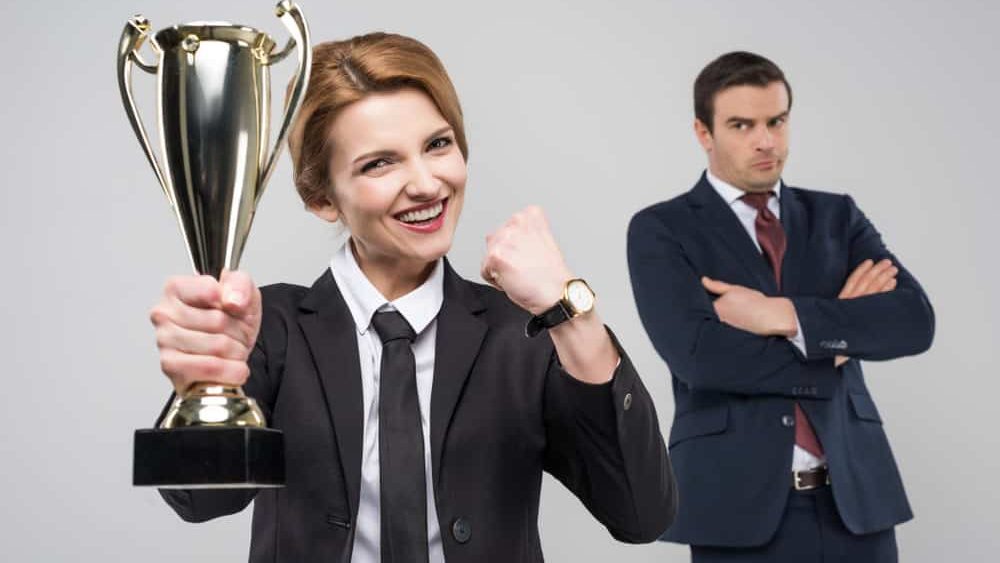 excited businesswoman with trophy cup and upset businessman behind, isolated on grey