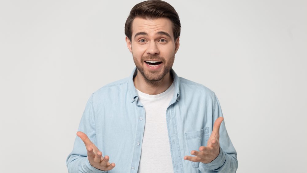Shocked young man in blue casual shirt isolated on grey studio background look at camera talking or telling news, surprised millennial guy make hand gesture announce good sale offer or discount