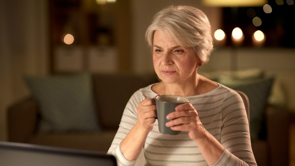 technology, old age and people concept - senior woman with laptop drinking coffee at home in evening