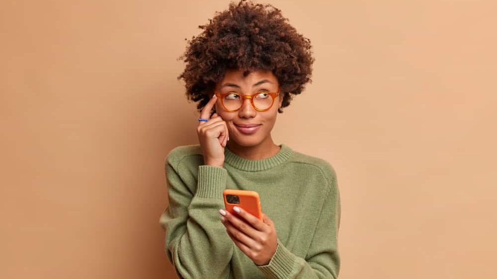 Pensive dark skinned young ethnic woman keeps index finger on temple tries to remember telephone number of someone holds modern smartphone and concentrated aside wears glasses casual jumper.