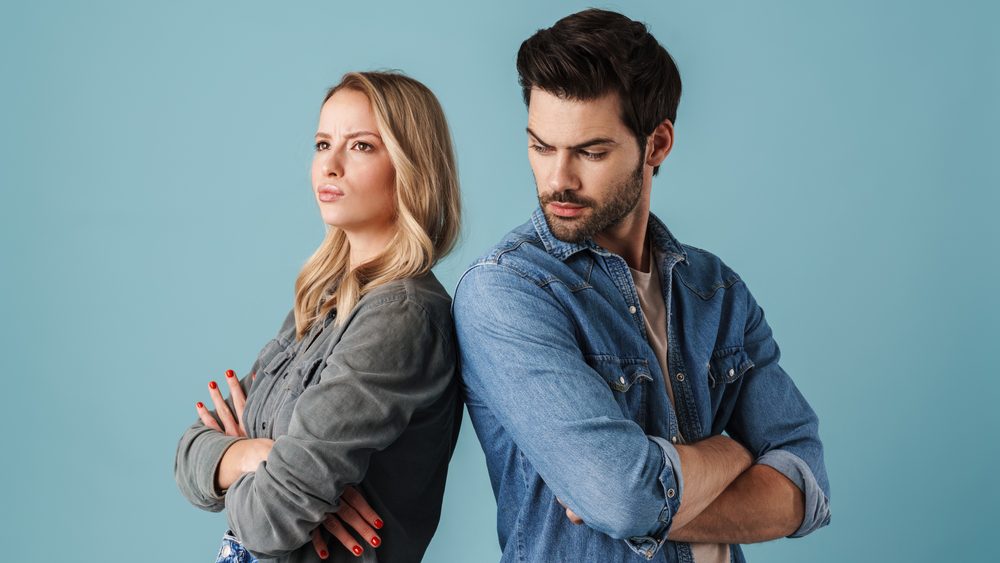 Young caucasian displeased man and woman standing back to back isolated over blue background