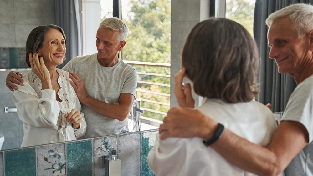 Senior woman before mirror touching her ear next to husband