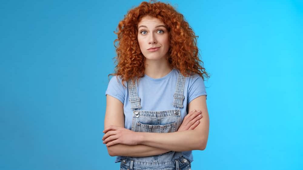 Unsure confused redhead curly woman look perplexed uncertain cross arms chest smirking stare camera full disbelief suspicious doubtful someone telling truth standing denim overalls blue background
