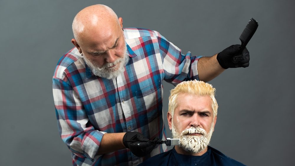 Bearded man coloring hair. Hair salon, hair coloring man. Attractive senior barber doing a haircut and haircolor for client at barber shop. Hipster guy dye his hair color on a gray background.