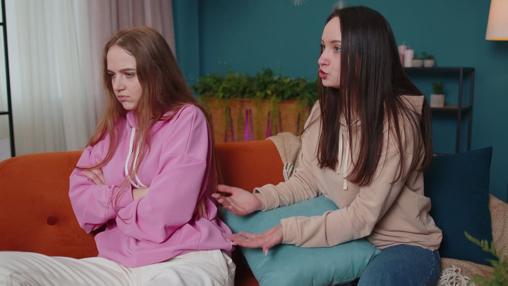 Girl sit separate from friend sibling turn back on sofa avoid talk after fight conflict, interracial upset. Two female women couple family have stress quarrel at home in living room. Woman apologizes