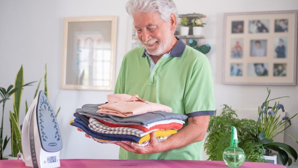 Smiling senior man helping with housework by ironing clothes on ironing board, looking proud to his ironed and folded shirts