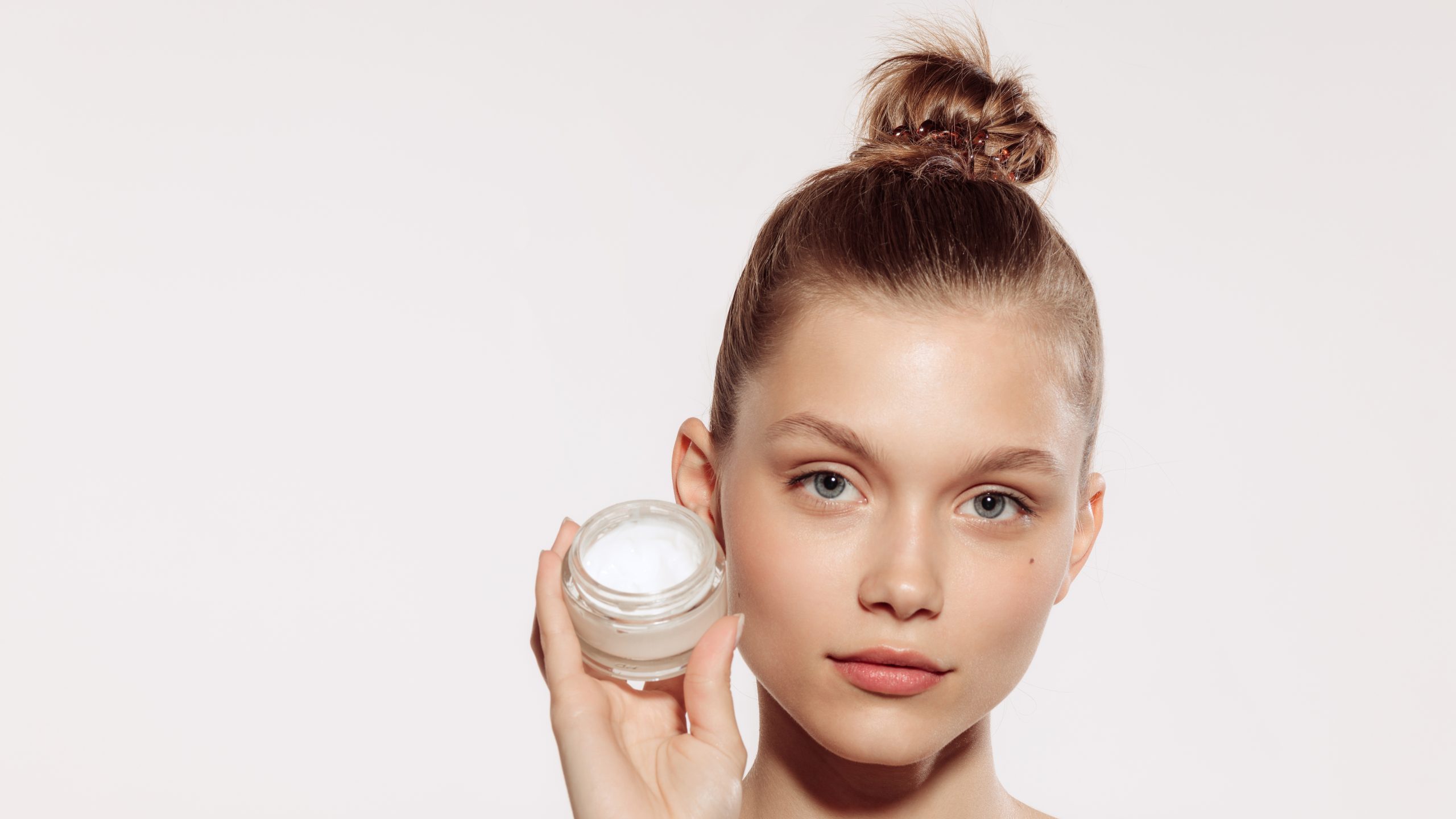 Half-length portrait of charming young girl without makeup holding cream bottle isolated over white studio background. Natural cosmetics treatment. Concept of beauty, skincare, cosmetology, spa