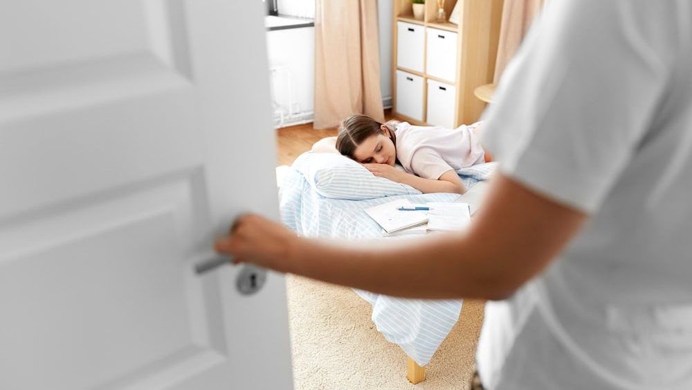 school, education and laziness concept - close up of parent opening door to kid's room and tired teenage student girl with notebooks sleeping on bed at home