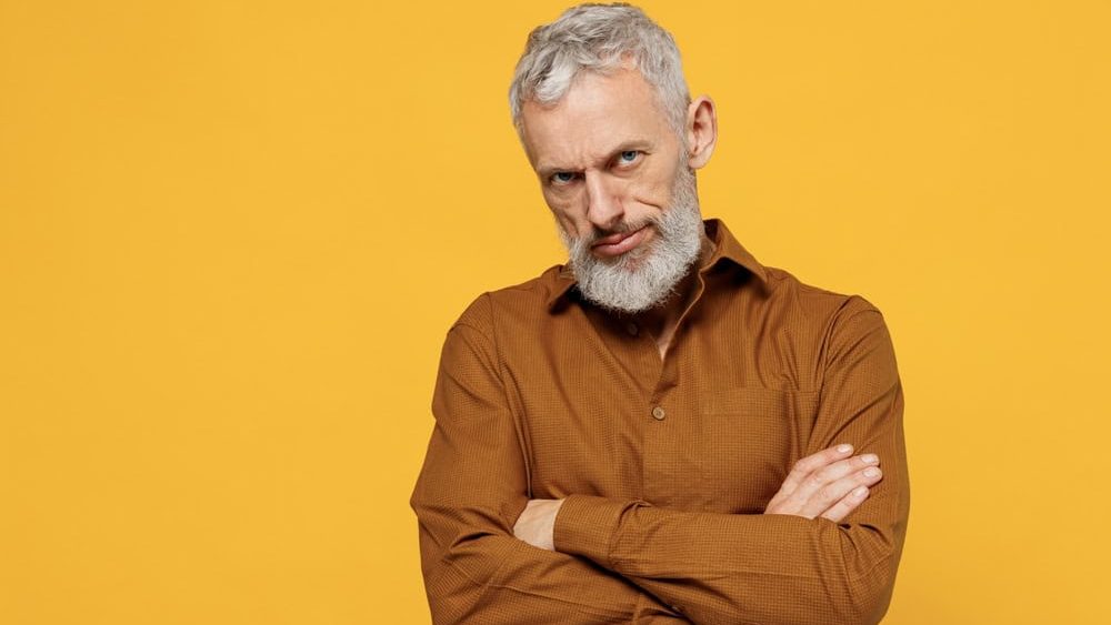 Angry frowning elderly gray-haired bearded man 40s years old wears brown shirt looking camera hold hands crossed isolated on plain yellow background studio portrait. People emotions lifestyle concept