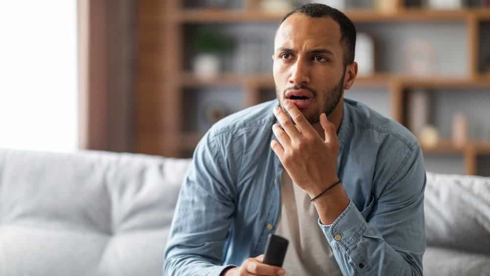 Shocked Black Man With Remote Controller In Hand Sitting On Couch At Home, Confused Young African American Guy Watching Tv Show In Living Room, Emotionally Reacting To Shocking Content, Copy Space