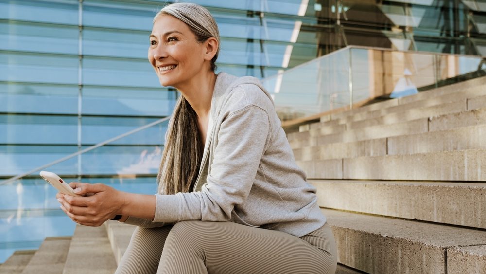 Asian mature woman in tracksuit using cellphone and sitting on stairs during workout outdoors