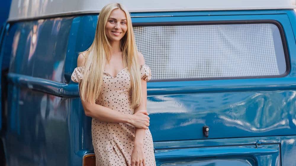 Attractive blonde caucasian female model posing outside against retro blue bus, toothy smiles. Vertical shot of Swedish beautiful girl at summertime in beige dress walking outside. Fashion model.