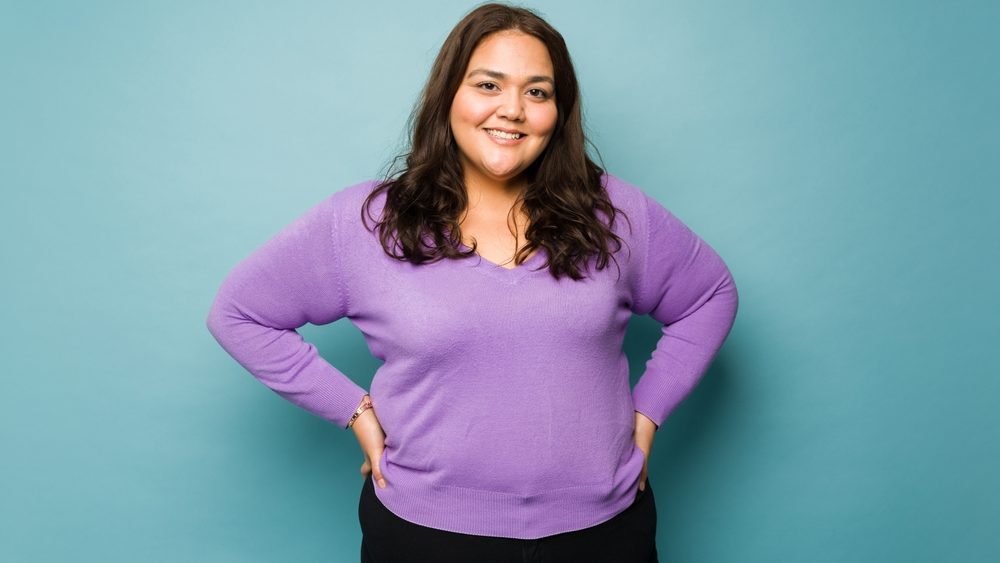 Cheerful latin fat woman with her hands on the hips and wearing casual clothes looking happy against a blue background